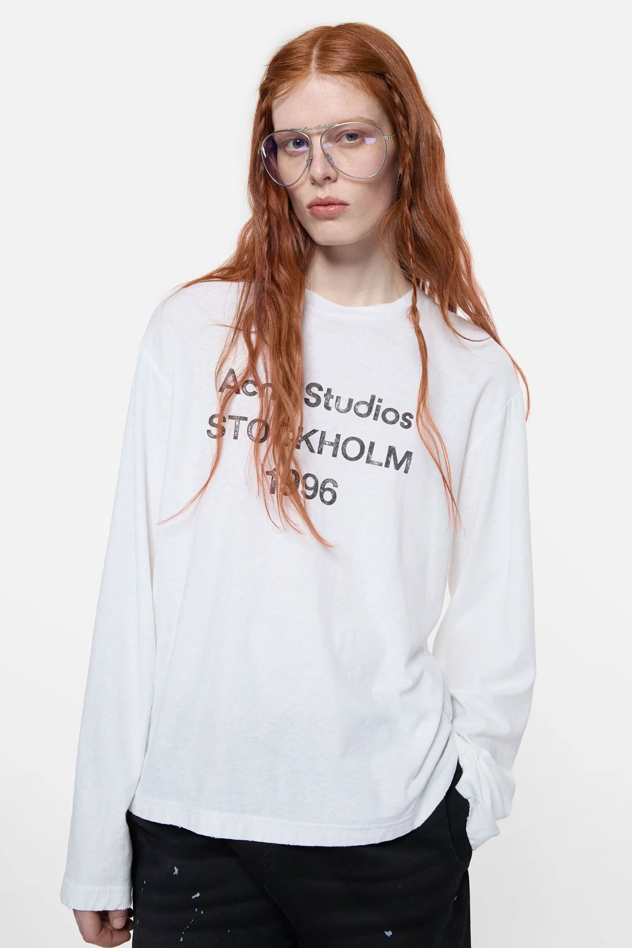 A female model wears a white long-sleeve t-shirt by Acne Studios with the signature 1996 logo on it and clear glasses.