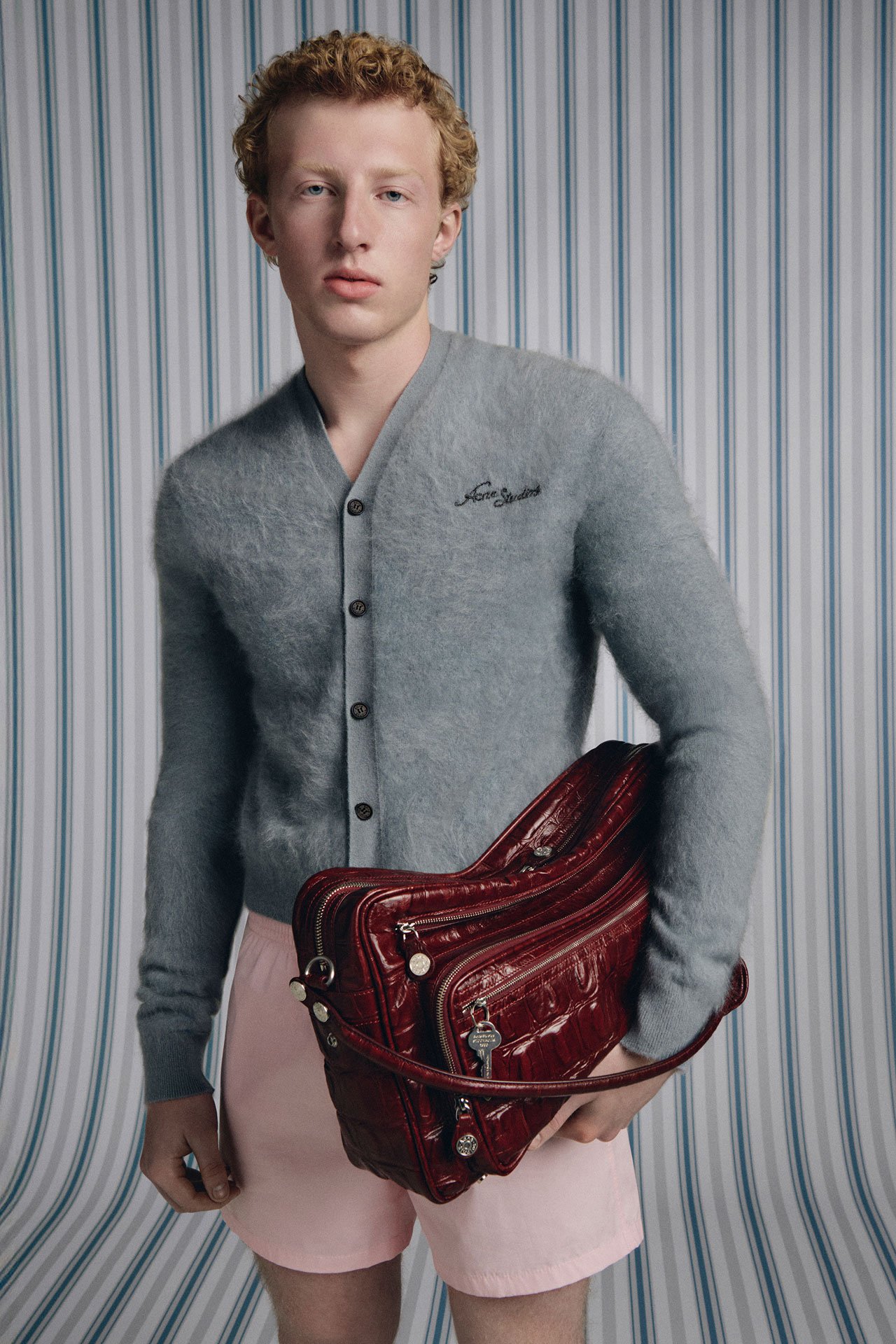 A male model wearing a white topless shirt, brown leather pants, black leather boots and holding a brown leather jacket.