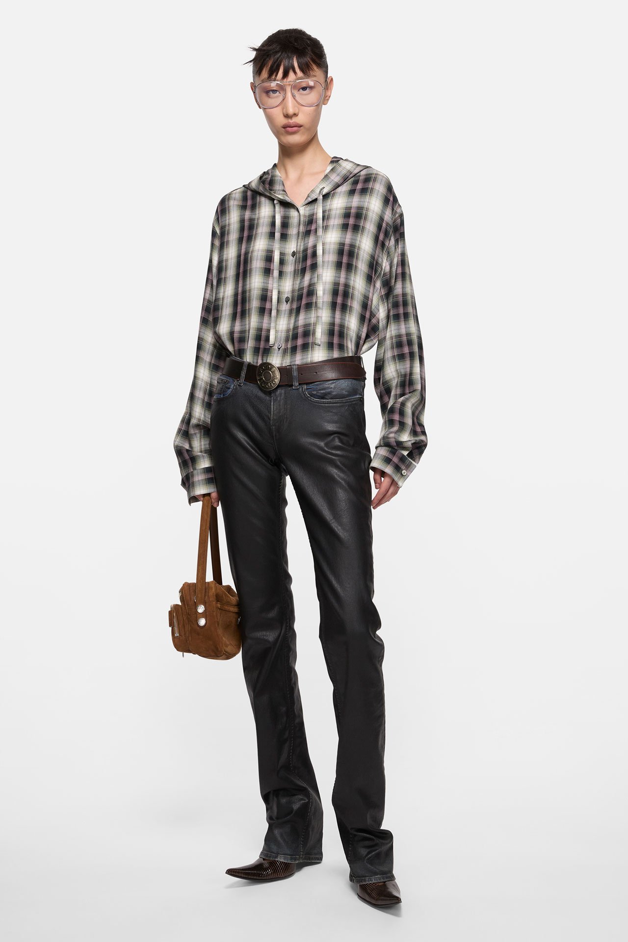 Female model showing Acne Studios new arrivals, with a checked beige-brown shirt, pressed black leather skinny jeans, holding a faux-leather brown Camero bag.