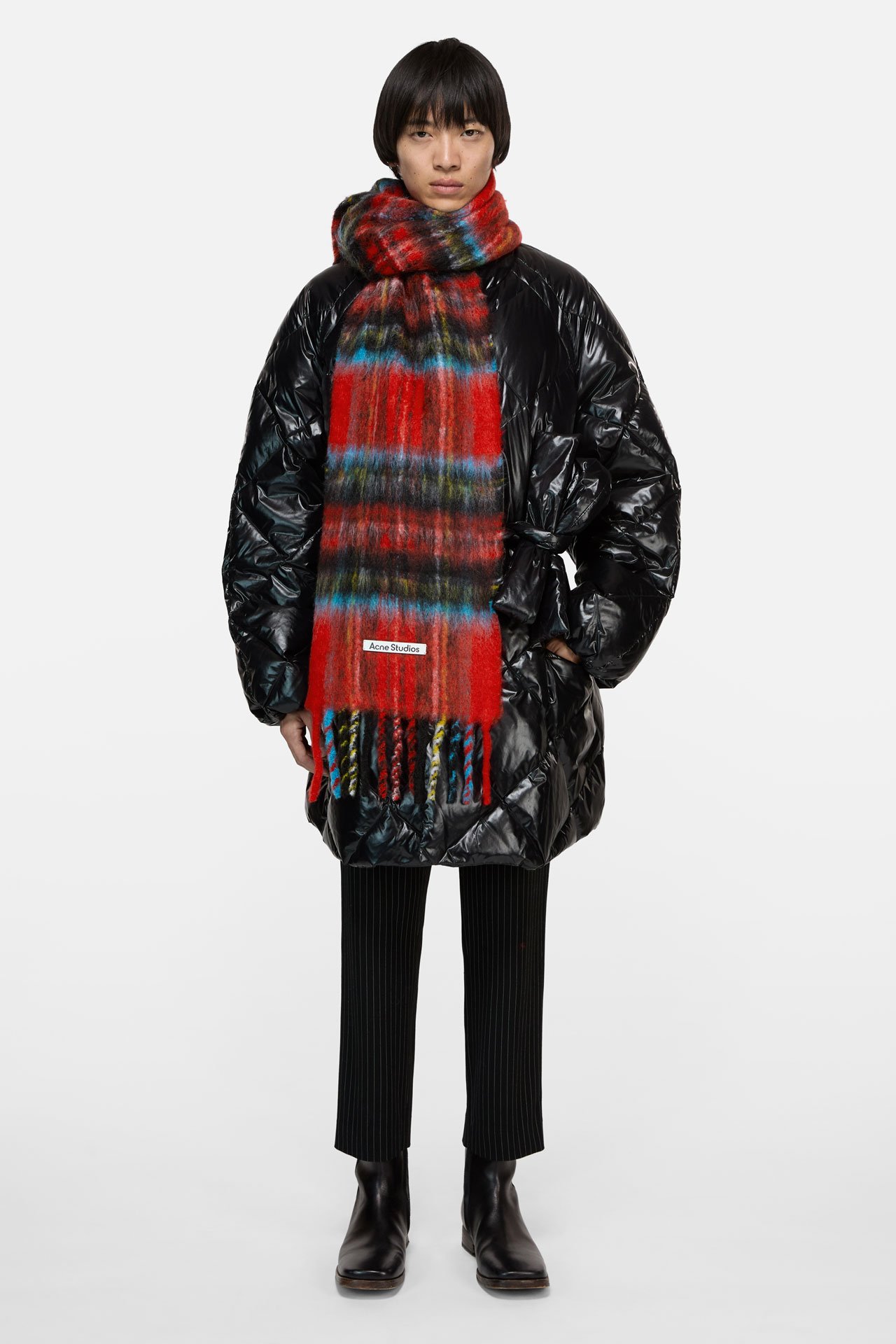 A dark haired male model wearing an Acne Studios red festive mohair check scarf, a big down blak winter coat, black pants and black boots.