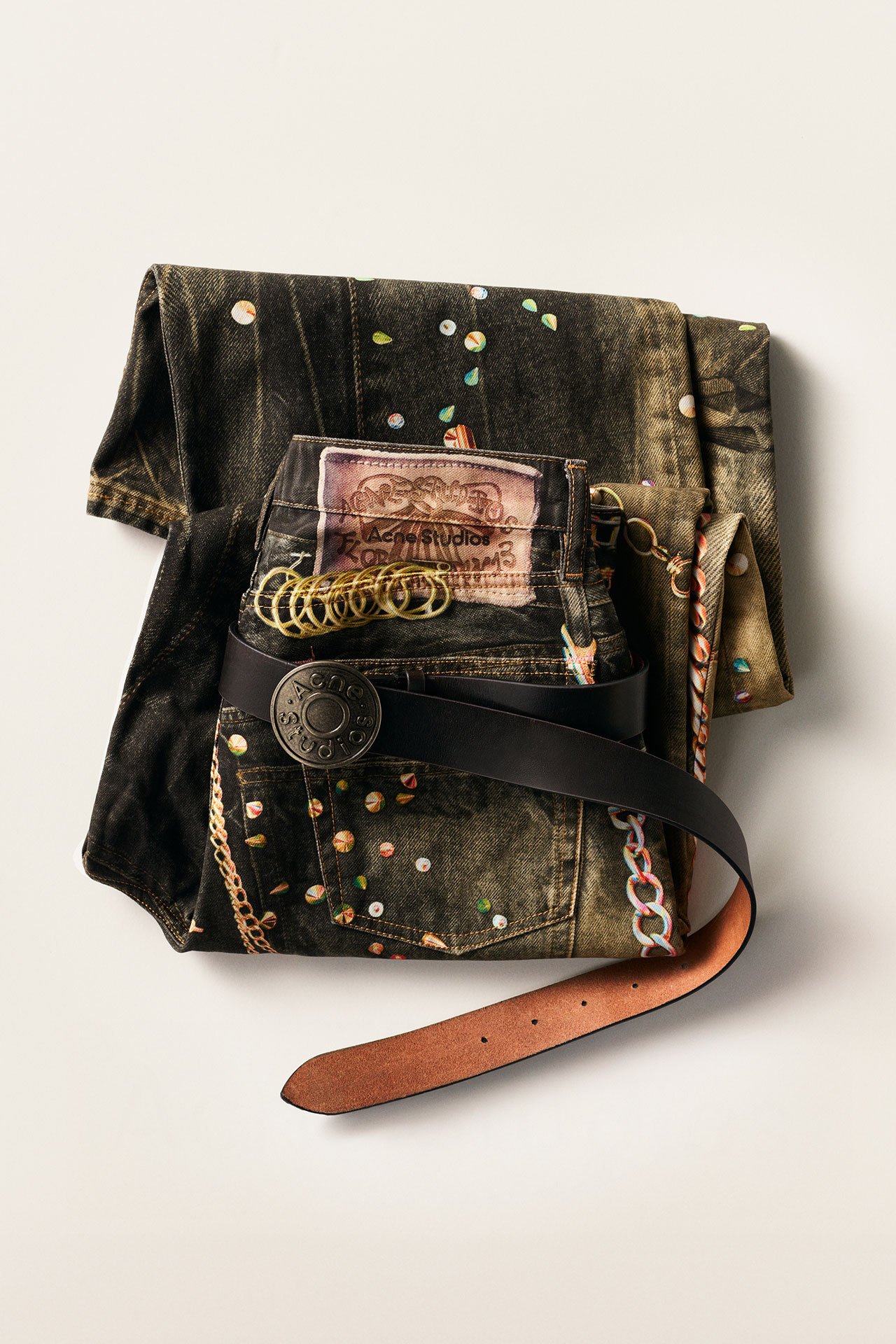Acne Studios SS26 Year of the Horse, a close-up of a folded dark jeans with green and beige wash with other patterns on it, with a black leather belt.