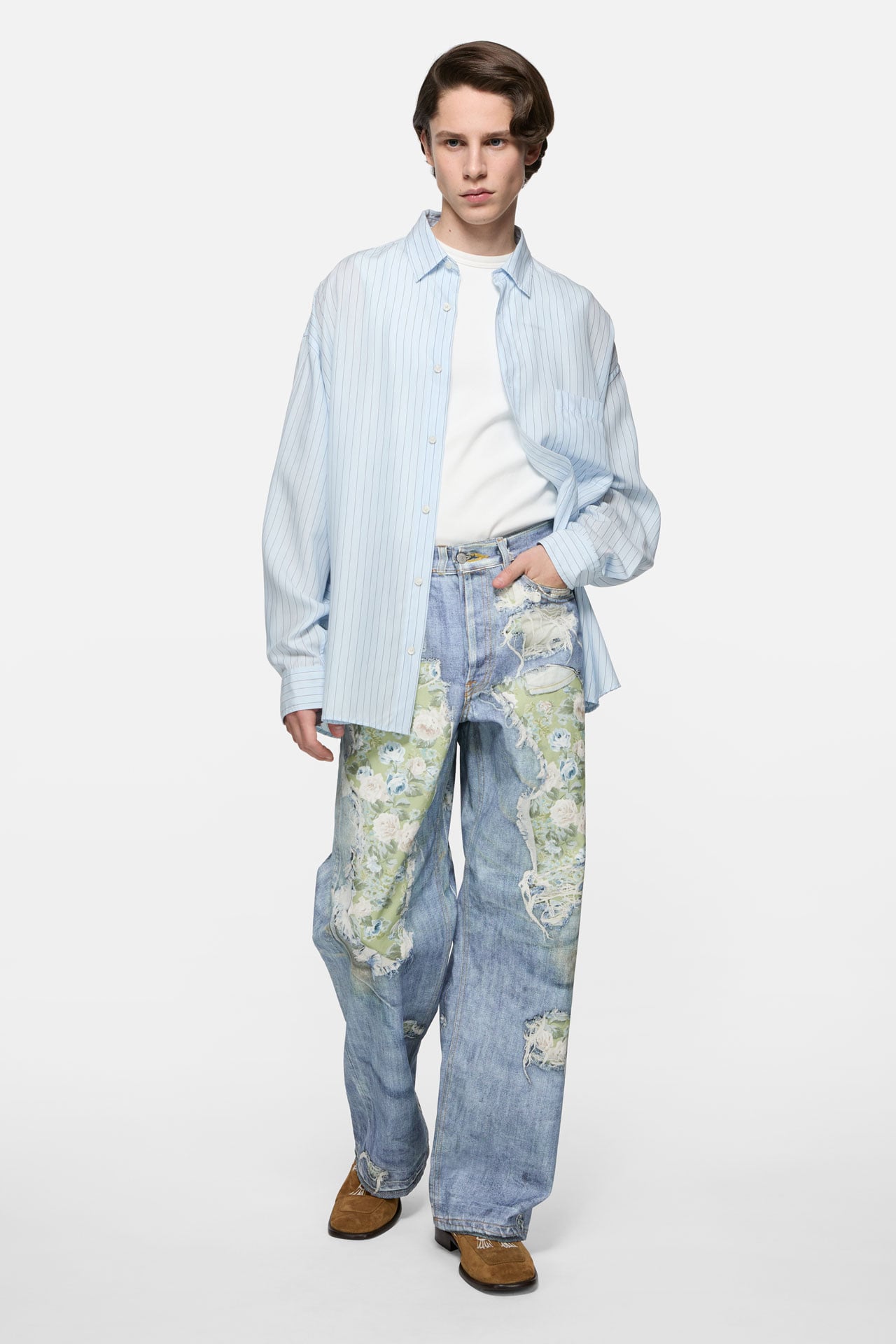 A male model wearing Acne Studios green-blue TLO jeans with rose patterns, white top and blue overshirt.