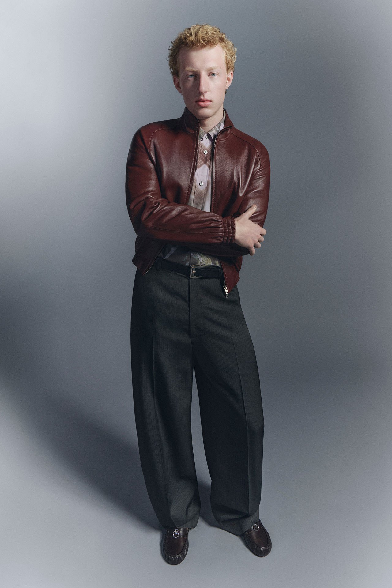 A male model wearing brown leather jacket, tailored black pants, black leather loafers and beige shirt.