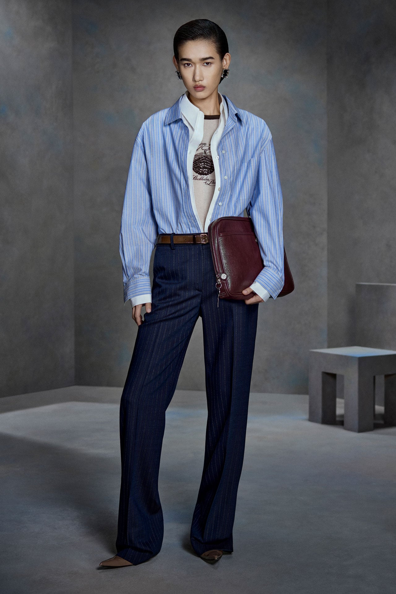 Acne Studios blue and white shirt open, beige tanktop with black motive under it, dark blue tailored pants and brown leather laptop case.