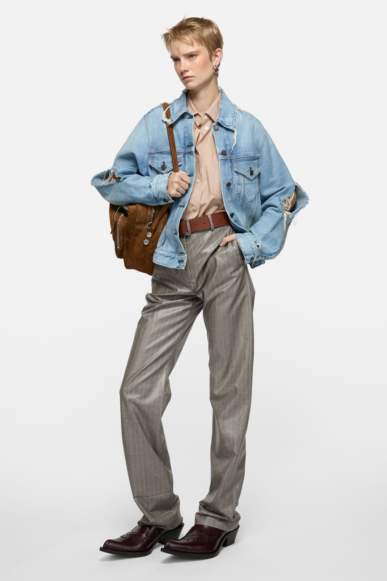 Female model wearing blue denim jacket, grey trousers, beige shirt, brown leather cowboy boots and holding a brown suede Camero bag.